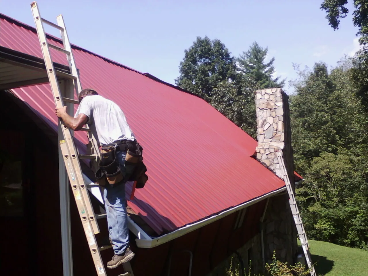 Roofer on a ladder beside a red metal roof and stone chimney — Soffit & Fascia Repair by Roofing Brothers in Lagunitas